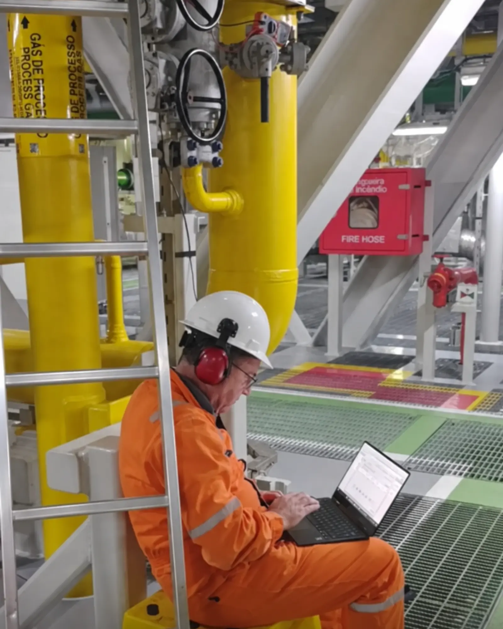 Técnico com EPI completo trabalha em notebook durante atendimento de campo em unidade industrial de gás de processo.